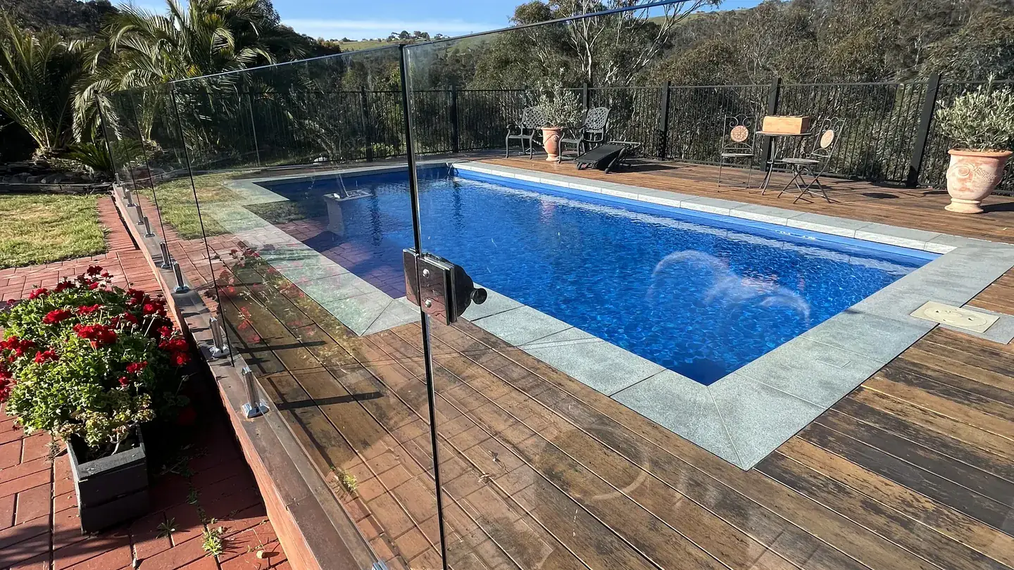 A swimming pool behind a frameless glass pool safety fence, with a timber deck and hills beyond
