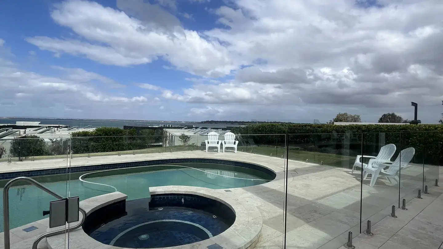A swimming pool behind a frameless glass pool safety fence on a hilltop property under a bright sky