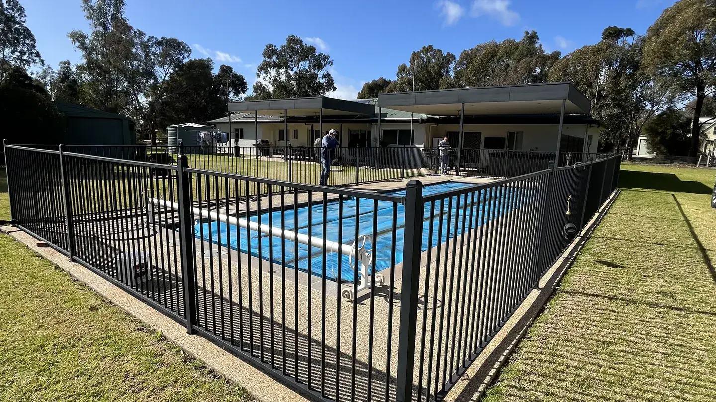 A backyard swimming pool behind a black aluminium pool safety fence, with a home and blue sky behind
