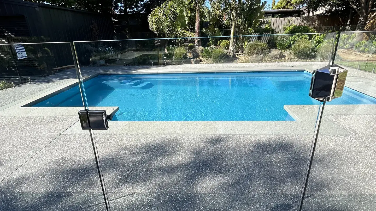 A swimming pool enclosed by a frameless glass pool safety fence, with gum trees and a clear blue sky