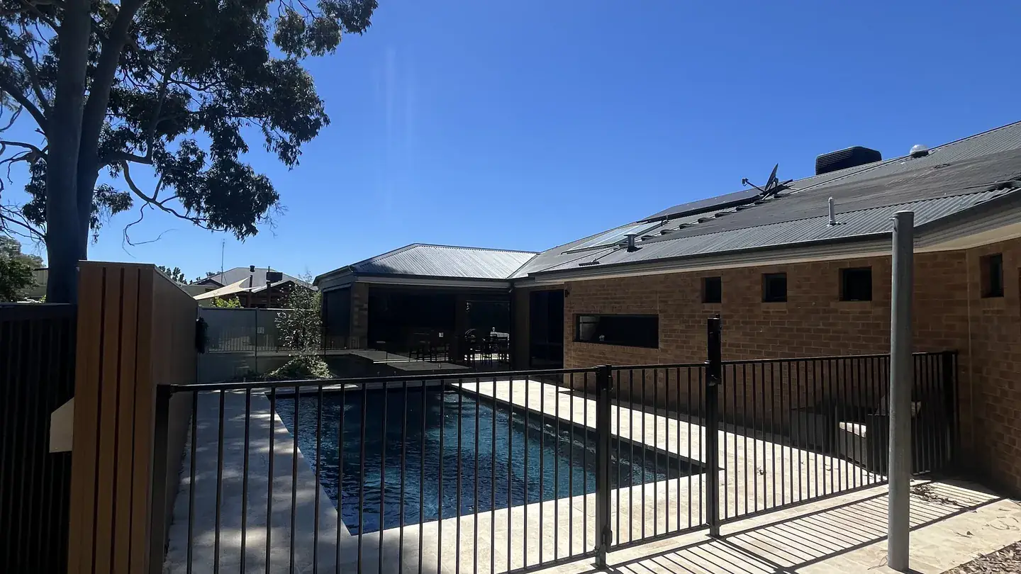A pool enclosed by a black aluminium pool safety fence, with a gum tree, home and bright sun
