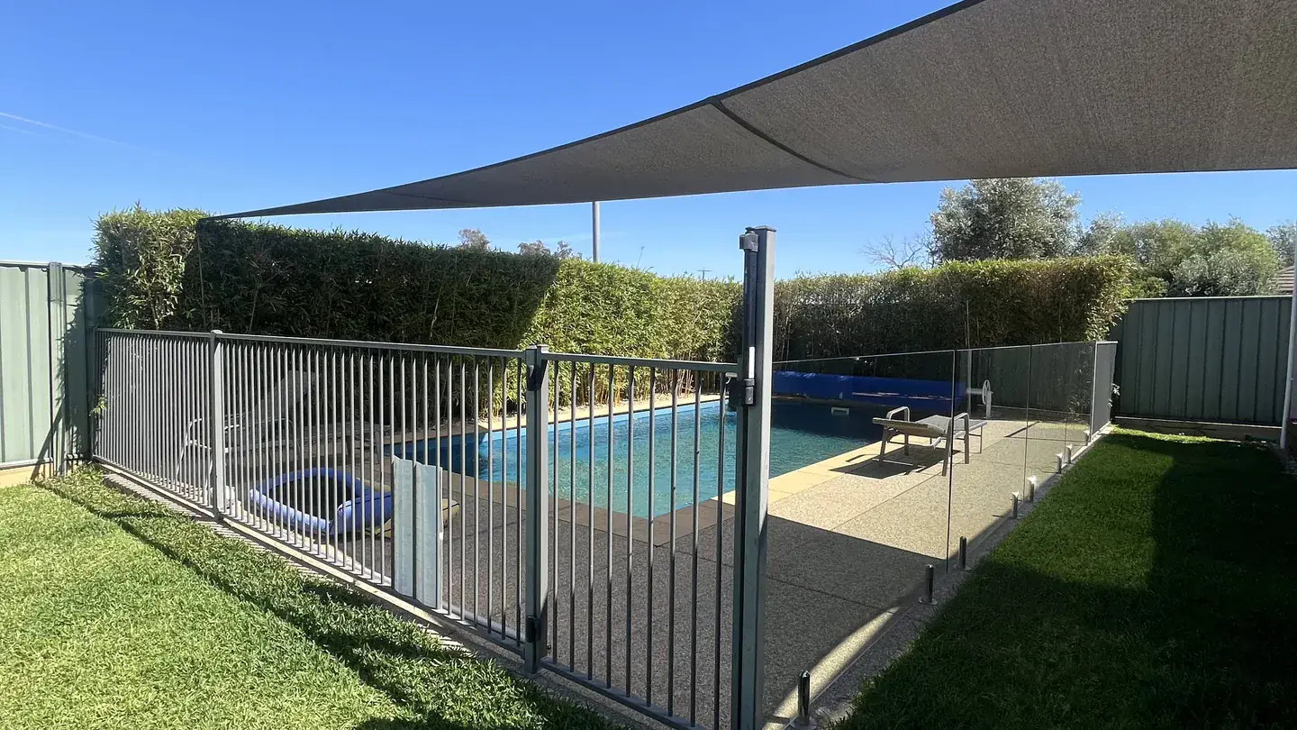 A black aluminium pool safety fence and self-latching gate beside a pool, with open lawn and a shade sail