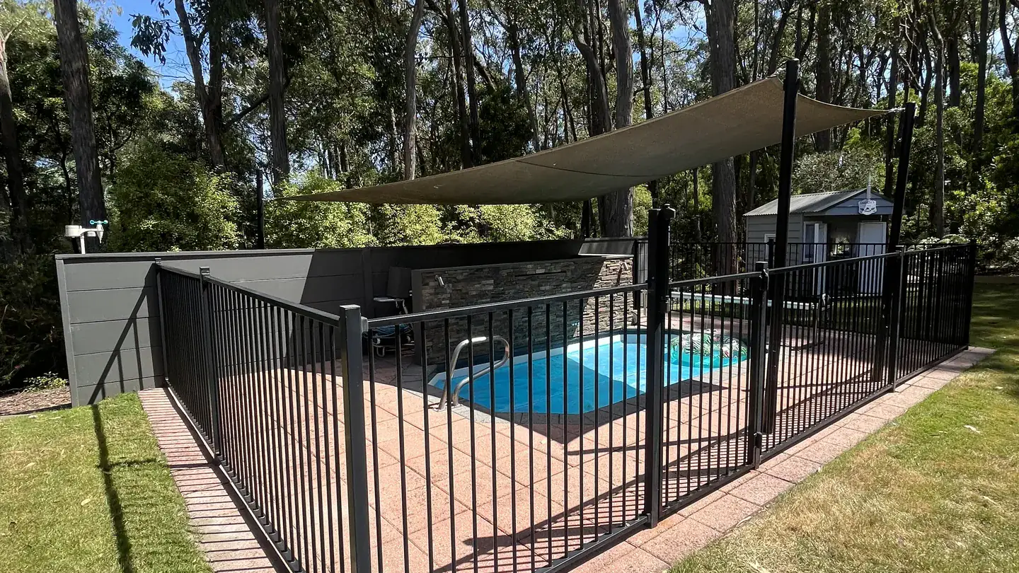 A backyard pool behind a black pool safety fence, with a shade sail and gum trees