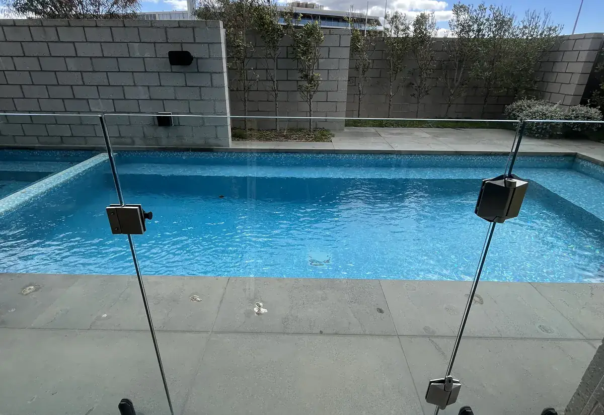 A frameless glass pool safety fence beside a swimming pool, against a rendered wall