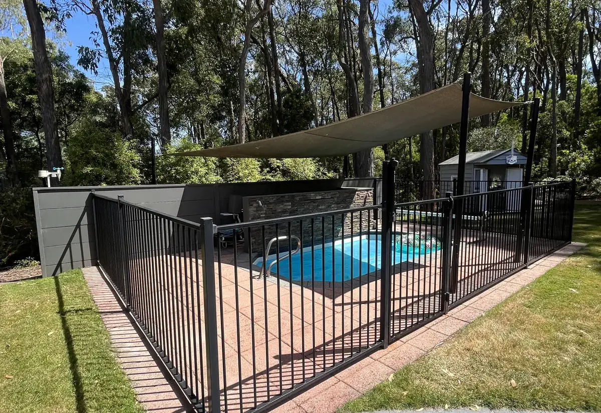 A backyard pool behind a black pool safety fence, with clear lawn, a shade sail and gum trees