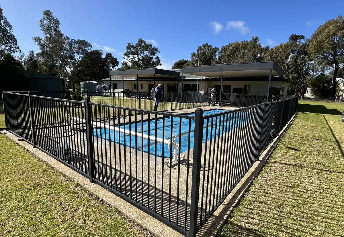 A swimming pool behind a black aluminium pool safety fence, with a home and blue sky behind